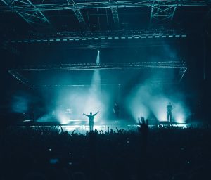Concert stage with a person standing in front spreading arms out wide, artifical smog in the background.